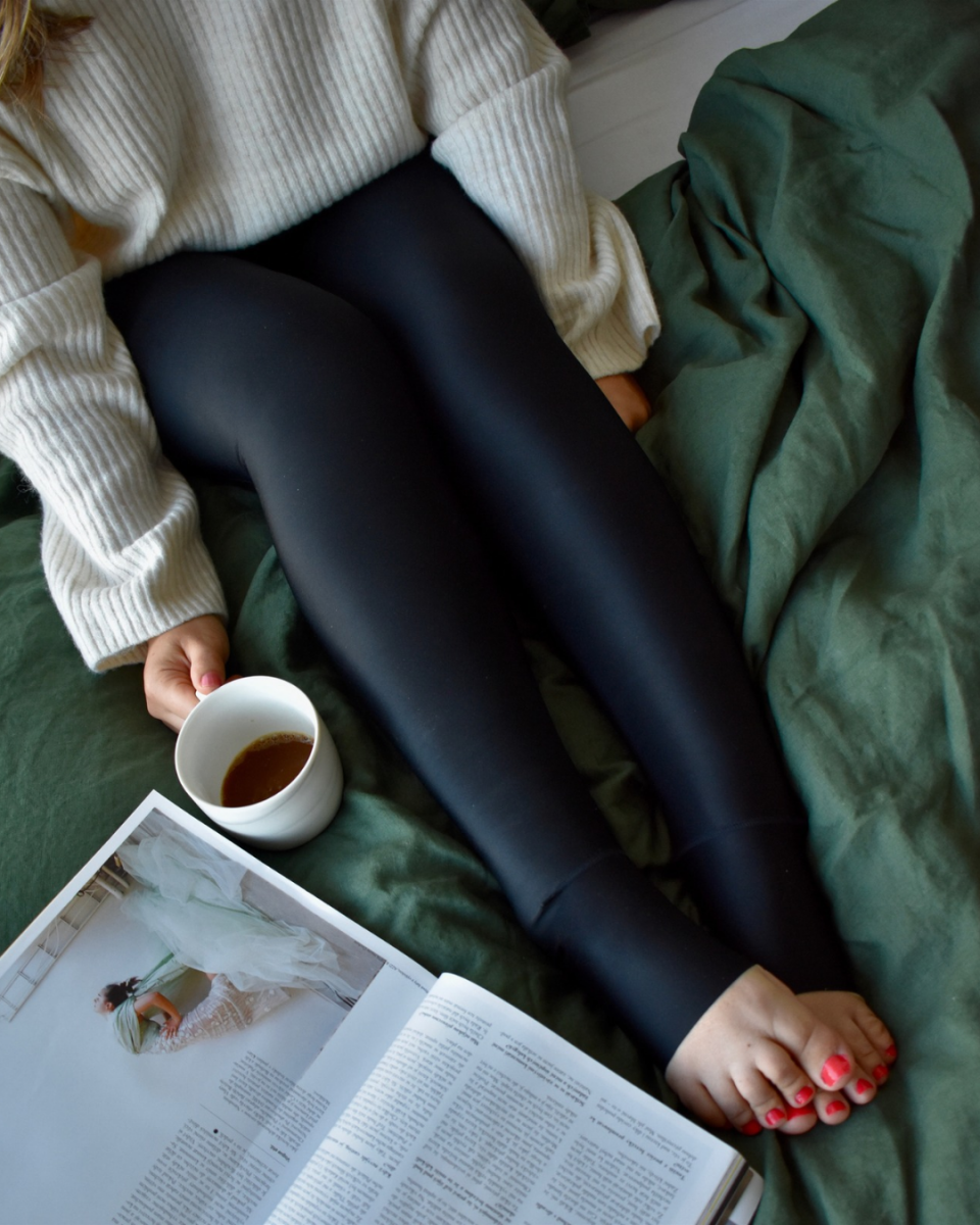 Eine Frau sitzt mit ihrem Kompressionsmieder auf der Couch. Neben ihr liegt ein Buch und sie hält eine Tasse Kaffee in der Hand.